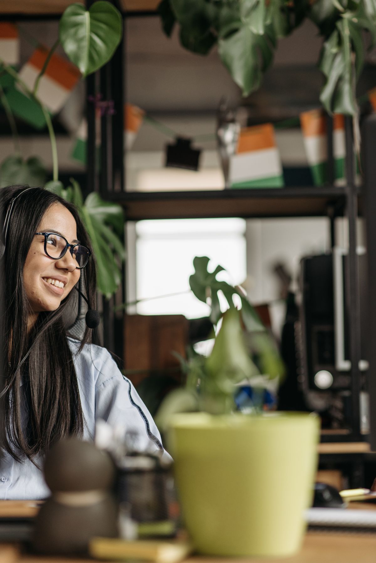 A woman working calmly at a home office desk surrounded by plants, wearing a headset